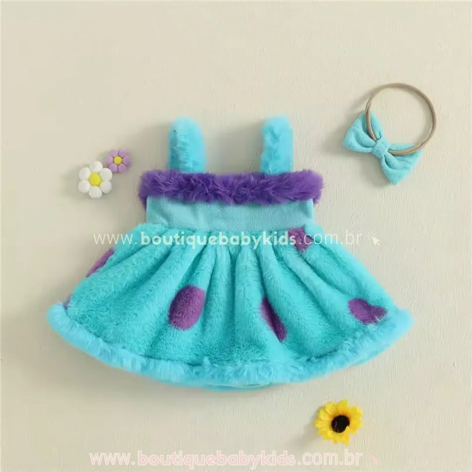 Vestido Sulley exposto em fundo claro, acompanhado de faixa com laço azul e flores decorativas ao lado