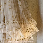 Close-up do tecido de tule bege com estampa de estrelas douradas brilhantes. Detalhe mostrando as camadas de babados na barra do Vestido Infantil Brilho das Estrelas.