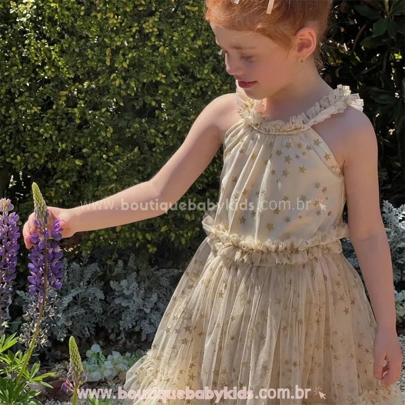 Menina usando vestido de festa infantil com estrelas, tocando em flores roxas. A imagem destaca o movimento leve do tecido de tule e o perfil do modelo frente única.
