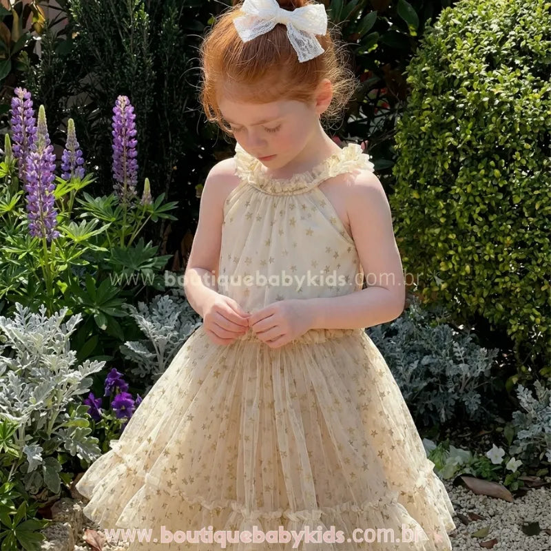 Menina vestindo o Vestido Infantil Brilho das Estrelas em um jardim. O vestido é bege champagne, rodado, com saia de tule em camadas e estampa de estrelas douradas.