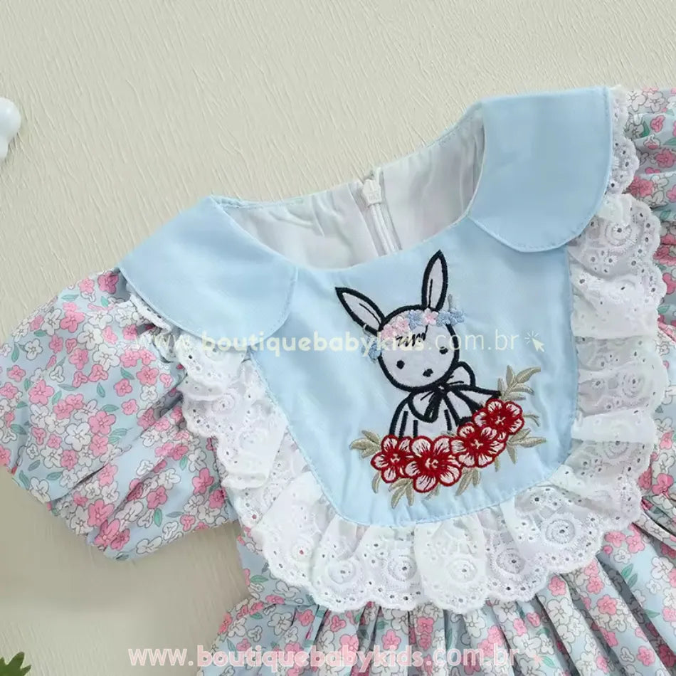 Detalhe do bordado central do vestido, apresentando um coelhinho delicado com laço floral na cabeça e flores vermelhas, cercado por babados de renda branca.