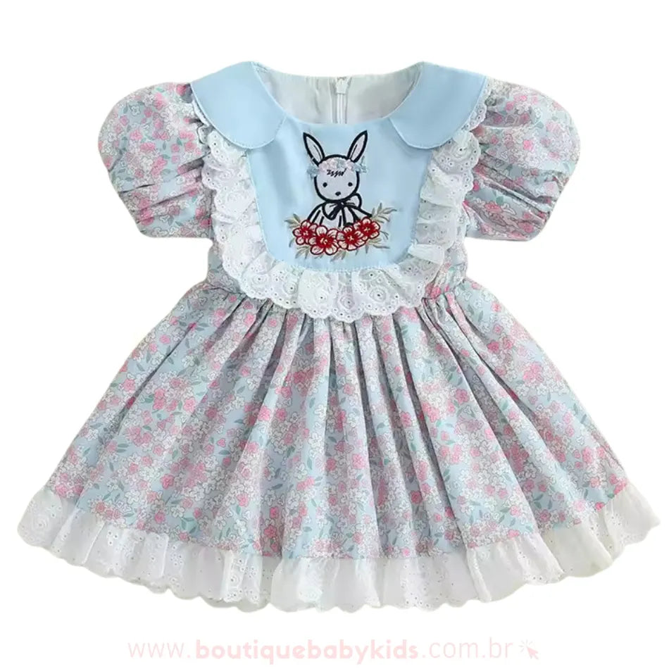 Vestido infantil azul floral com detalhes bordados de coelhinho e flores na parte frontal, gola arredondada azul clara e babados de renda branca ao redor do busto.