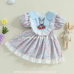 Vestido infantil azul floral com manga bufante e saia rodada, decorado com bordado de coelho com flores vermelhas e babados de renda na parte frontal.